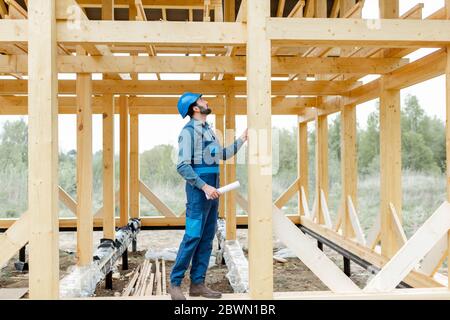 Costruttore in tute blu e elmetto sul cantiere, costruzione di case con telaio in legno Foto Stock