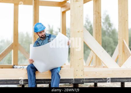 Costruttore in tute blu e cappello con blueprint sul cantiere. Costruzione di case di legno con struttura Foto Stock