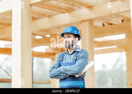 Ritratto di un bel costruttore di cofidenti in tute blu e cappello sul cantiere. Costruzione di case di legno con struttura Foto Stock
