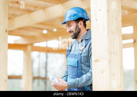 Ritratto di un bel costruttore di cofidenti in tute blu e cappello sul cantiere. Costruzione di case di legno con struttura Foto Stock