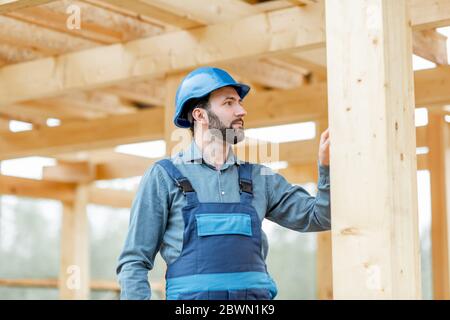 Ritratto di un bel costruttore di cofidenti in tute blu e cappello sul cantiere. Costruzione di case di legno con struttura Foto Stock