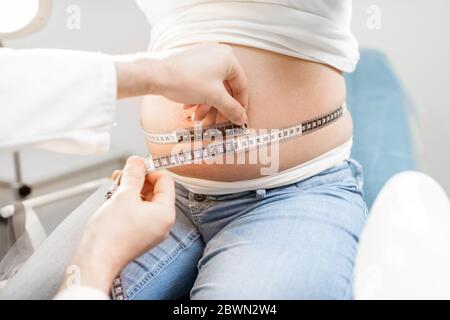 Medico che misura la pancia della donna incinta con un nastro durante un esame medico, vista ritagliata senza volto messo a fuoco sull'addome Foto Stock