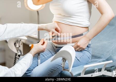 Medico che misura la pancia della donna incinta con un nastro durante un esame medico, vista ritagliata senza volto messo a fuoco sull'addome Foto Stock