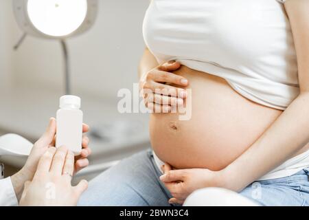 Medico che tiene alcuni farmaci o integratori per una donna incinta durante una consultazione, vista ravvicinata focalizzata sulla pancia incinta. Concetto di farmaco per le donne incinte Foto Stock