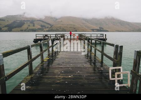 Donna su una giacca rossa in piedi a Port Levy Jetty, Banks Peninsula, Nuova Zelanda Foto Stock