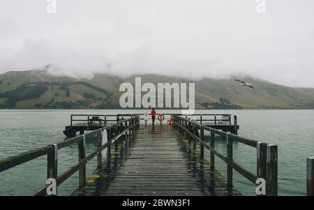 Donna su una giacca rossa in piedi a Port Levy Jetty, Banks Peninsula, Nuova Zelanda Foto Stock