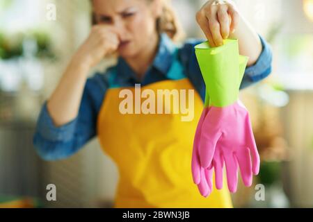 Primo piano su donna disgustata in grembiule arancione e guanti di gomma rosa nel soggiorno in giornata di sole con guanti puzzolenti. Foto Stock