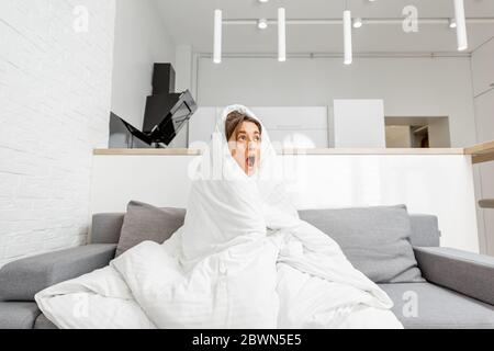 Donna sonnolenta coperta da una coperta bianca sul divano di casa, ampia vista interna. Concetto di auto-isolamento e malattie influenzali a casa Foto Stock