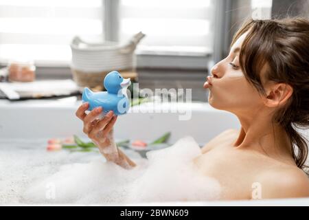 Giovane e allegra donna che prende un bagno, sdraiata nella vasca da bagno con anatra in gomma, rilassante nel bagno a casa Foto Stock