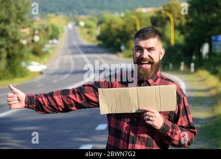 Hipster cercare di fermare l'auto con cartoncino e pollice in su gesto, spazio di copia. Uomo con viso e barba allegri che viaggia con un'escursione con strada sullo sfondo. Concetto di viaggio e di escursionismo. Foto Stock