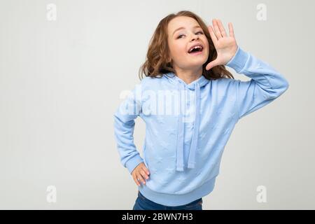 L'affascinante ragazza europea in una felpa con cappuccio blu racconta le notizie mentre tiene le mani in bocca su uno sfondo chiaro Foto Stock