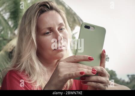 Donna bionda adulta che tiene, usando e guardando lo smartphone all'aperto sull'isola di Bali, Indonesia. Foto Stock
