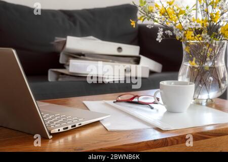 Laptop, documenti e raccoglitori di anelli su un tavolo di legno e il divano dietro, lavorando in un ufficio temporaneo durante la pandemia coronavirus cr Foto Stock