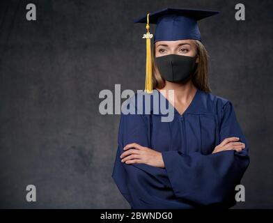 Giovane donna laureata in maschera nera con le mani incrociate celebrando il suo grado universitario indossando cappuccio blu e abito guardando al lato Foto Stock