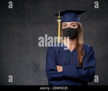 Giovane donna laureata in maschera nera con le mani incrociate celebrando il suo grado universitario indossando il cappuccio blu e abito guardando dritto Foto Stock