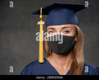 Giovane donna laureata in maschera nera che celebra il suo grado universitario indossando cappello blu e abito da vicino Foto Stock