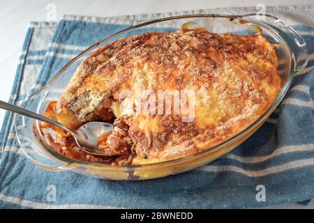 Moussaka cotta in casseruola di vetro, piatto tradizionale greco di melanzane, patate, carne tritata e pomodori su un asciugamano blu da cucina, fuoco selezionato, Foto Stock