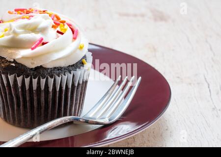 primo piano di una cupcake al cioccolato in un rivestimento di carta con glassa di vaniglia con spruzzini rosa e giallo su un piatto rosso e bianco con forchetta Foto Stock