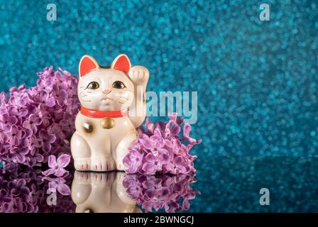 Simbolo giapponese di buona fortuna gatto neko maneki incorniciato da fiori lilla su uno sfondo blu bokeh con riflessione Foto Stock