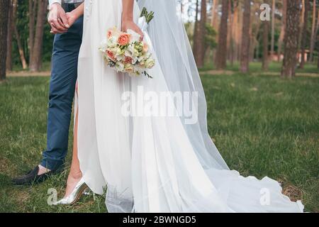 una sposa in un bel vestito con un treno che tiene un bouquet di fiori e verde Foto Stock