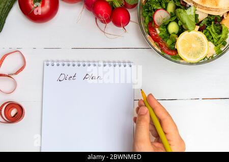Donna mano che tiene la penna e le parole che scrivono programma di dieta in un blocco note su tavola di legno bianco con la ciotola di insalata e verdure Foto Stock