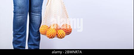 Giovane donna in jeans blu è su tiene borsa stringa con vari agrumi su sfondo grigio. Concetto zero sprechi, eco-compatibile. Spazio di copia Foto Stock