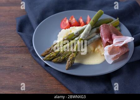 Asparagi verdi cotti su purè di patate con salsa di hollandaise chiara, prosciutto e pomodori serviti su un piatto blu e tovagliolo su una rustica linguetta di legno scuro Foto Stock