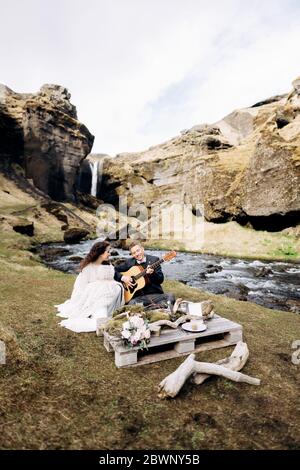 Una coppia di nozze è seduta sulla riva di un fiume di montagna, ad un tavolo per una cena di nozze. Lo sposo suona e canta per la sposa. Destinazione Foto Stock