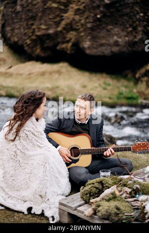 Una coppia di nozze è seduta sulla riva di un fiume di montagna, ad un tavolo per una cena di nozze. Lo sposo suona e canta per la sposa. Destinazione Foto Stock