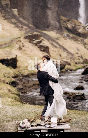 Destinazione Islanda matrimonio, vicino alla cascata Kvernufoss. Matrimonio in coppia sulla riva di un fiume di montagna. Lo sposo indossa la sposa in una coperta di lana Foto Stock