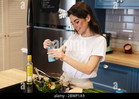 Bella giovane bruna donna che cucina insalata mentre si è in piedi al banco della cucina, peppering Foto Stock