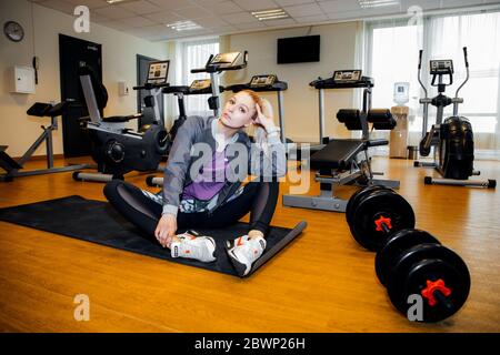 Bella ragazza in forma si siede in una sala fitness con vari attrezzi fitness intorno a lei, manubri nella parte anteriore; ha i suoi capelli in una coda e indossa un vestito sportivo Foto Stock