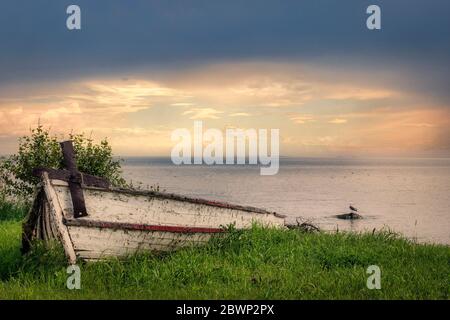 immagine orizzontale di un vecchio cinghiale a file bianche con vernice che si stacca sull'erba verde accanto a un tranquillo lago blu sotto un bel sole che tramonta Foto Stock