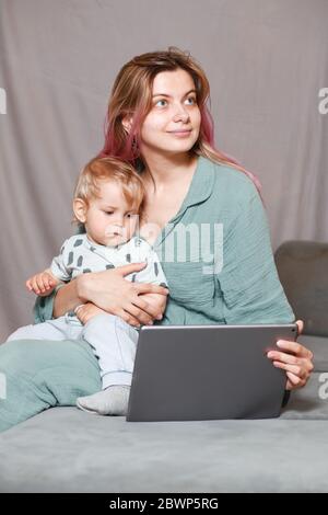 Rimani a casa, mamma lavora in remoto su un computer portatile, prendendosi cura del bambino. Una giovane madre in congedo di maternità sta cercando di lavorare come freelance w Foto Stock