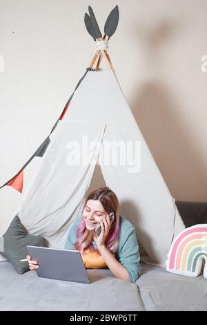 Una giovane madre lavora su un computer tablet a casa, in una capanna per bambini fatta di lenzuola. Concetto di quarantena durante una pandemia Foto Stock
