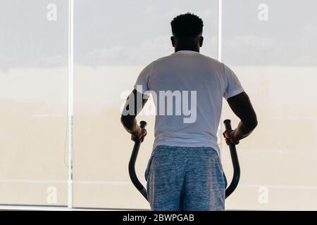 Vista posteriore di uomo afroamericano in forma sportiva facendo passi su macchina ellittica durante l'allenamento in palestra moderna. Foto Stock