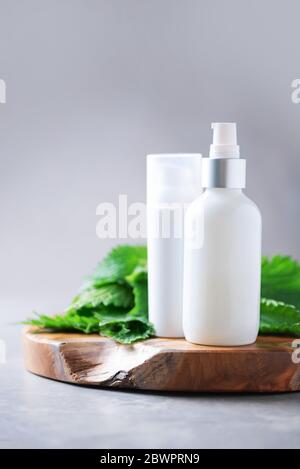 Centro benessere naturale, prodotti cosmetici a base di erbe. Crema di ortica, crema, shampoo o sapone in bottiglia bianca e foglie di bollitori fresche su sfondo grigio. Erba medicinale per h. Foto Stock