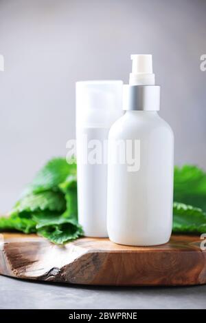 Crema di ortica, crema, shampoo o sapone in bottiglia bianca e foglie di bollitori fresche su sfondo grigio. Erbe medicinali per la salute e la bellezza, la cura della pelle e. Foto Stock