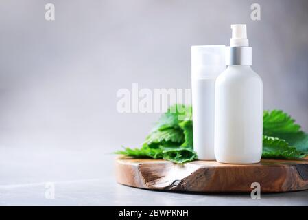Centro benessere naturale, prodotti cosmetici a base di erbe. Crema di ortica, crema, shampoo o sapone in bottiglia bianca e foglie di bollitori fresche su sfondo grigio. Erba medicinale per h. Foto Stock