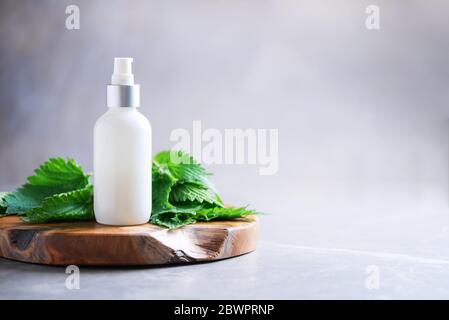 Crema di ortica, crema, shampoo o sapone in bottiglia bianca e foglie di bollitori fresche su sfondo grigio. Erbe medicinali per la salute e la bellezza, la cura della pelle e. Foto Stock