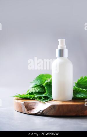 Centro benessere naturale, prodotti cosmetici a base di erbe. Crema di ortica, crema, shampoo o sapone in bottiglia bianca e foglie di bollitori fresche su sfondo grigio. Erba medicinale per h. Foto Stock