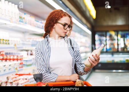 Giovane donna pensiva in occhiali e camicia a righe con carrello di shopping pensieroso guardando su bottiglia di latte nel supermercato Foto Stock