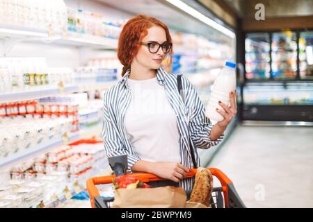 Bella ragazza in occhiali e camicia a righe con carrello pieno di prodotti che guarda pensierosamente su bottiglia di latte in mani in supermercato Foto Stock