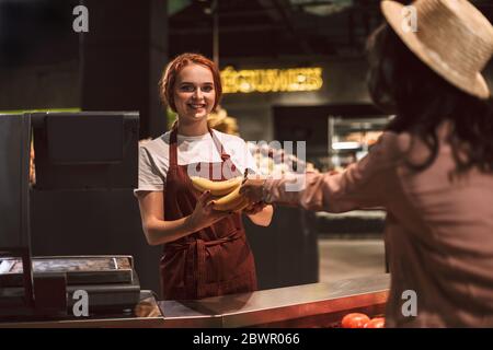Giovane venditore sorridente in grembiule dietro il contatore che guarda felicemente in macchina fotografica mentre dà banane al cliente nel supermercato moderno Foto Stock