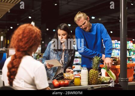 Giovane donna con lista della spesa in piedi vicino alla scrivania del cassiere mentre l'uomo che parla sul cellulare con i prodotti nel supermercato Foto Stock