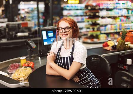 Cassa sorridente graziosa in occhiali e grembiule a righe che guarda felicemente in macchina fotografica mentre lavora in un supermercato moderno Foto Stock