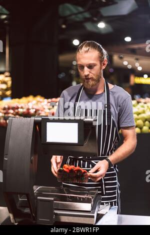 Venditore giovane in grembiule che pesa fragole su scala in supermercato Foto Stock