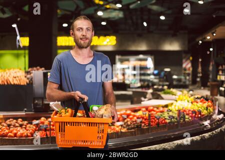 Giovane uomo sorridente sognando in macchina fotografica che tiene il cestino pieno di prodotti in mano nel supermercato Foto Stock