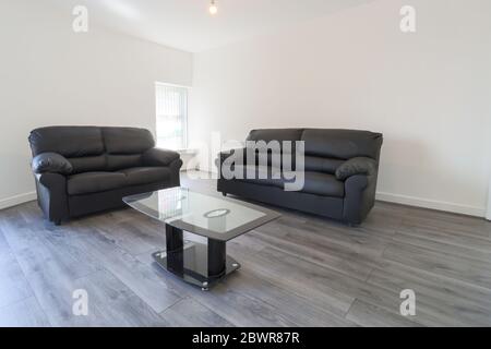Un moderno soggiorno britannico con due divani in pelle nera su un pavimento in legno grigio con pareti bianche e moderne persiane alla finestra Foto Stock