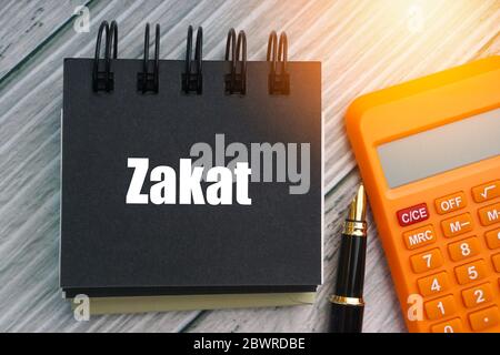 TESTO DI ZAKAT o DELLA TASSA ISLAMICA con penna stilografica, calcolatrice e blocco note su sfondo di legno. Zakat è una forma di elemosina trattata nell'Islam come religi Foto Stock